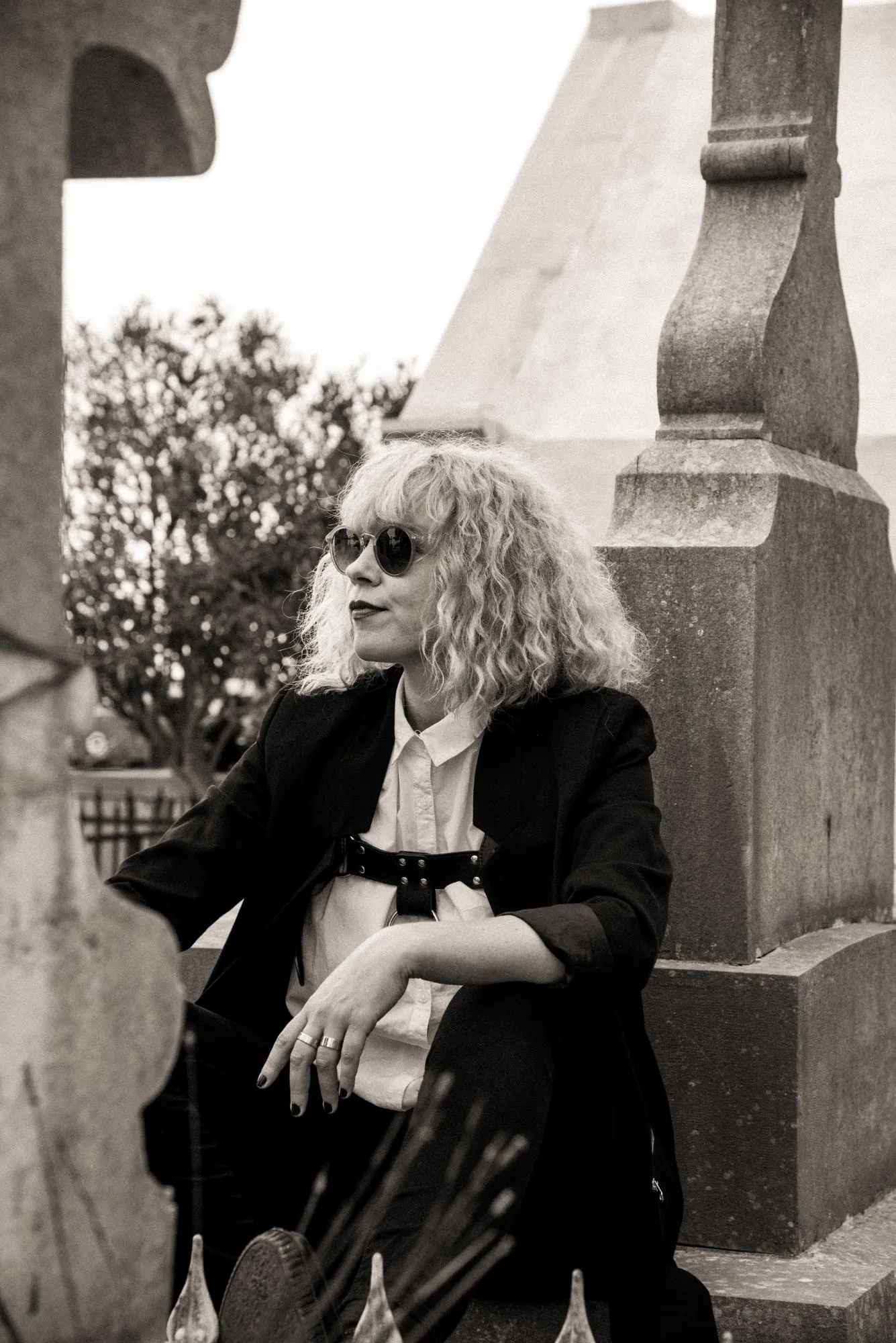 Emeraldia sitting between stone graves, sunglasses, black jacket and a harness over a white shirt