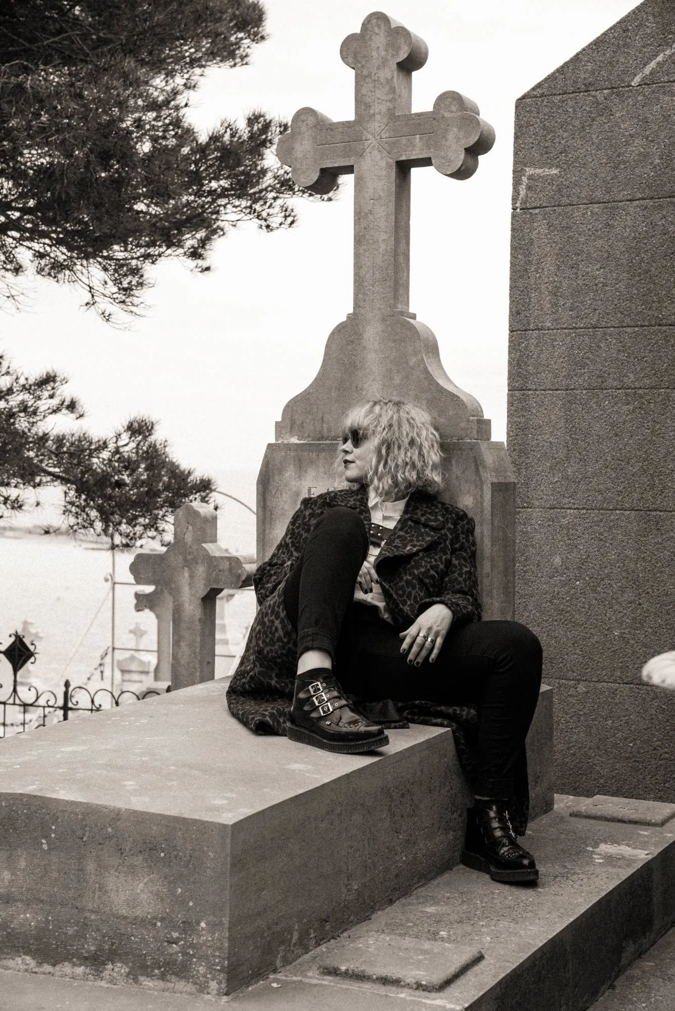 Emeraldia sitting on a grave at the foot of a stone cross, leopard coat and buckled boots