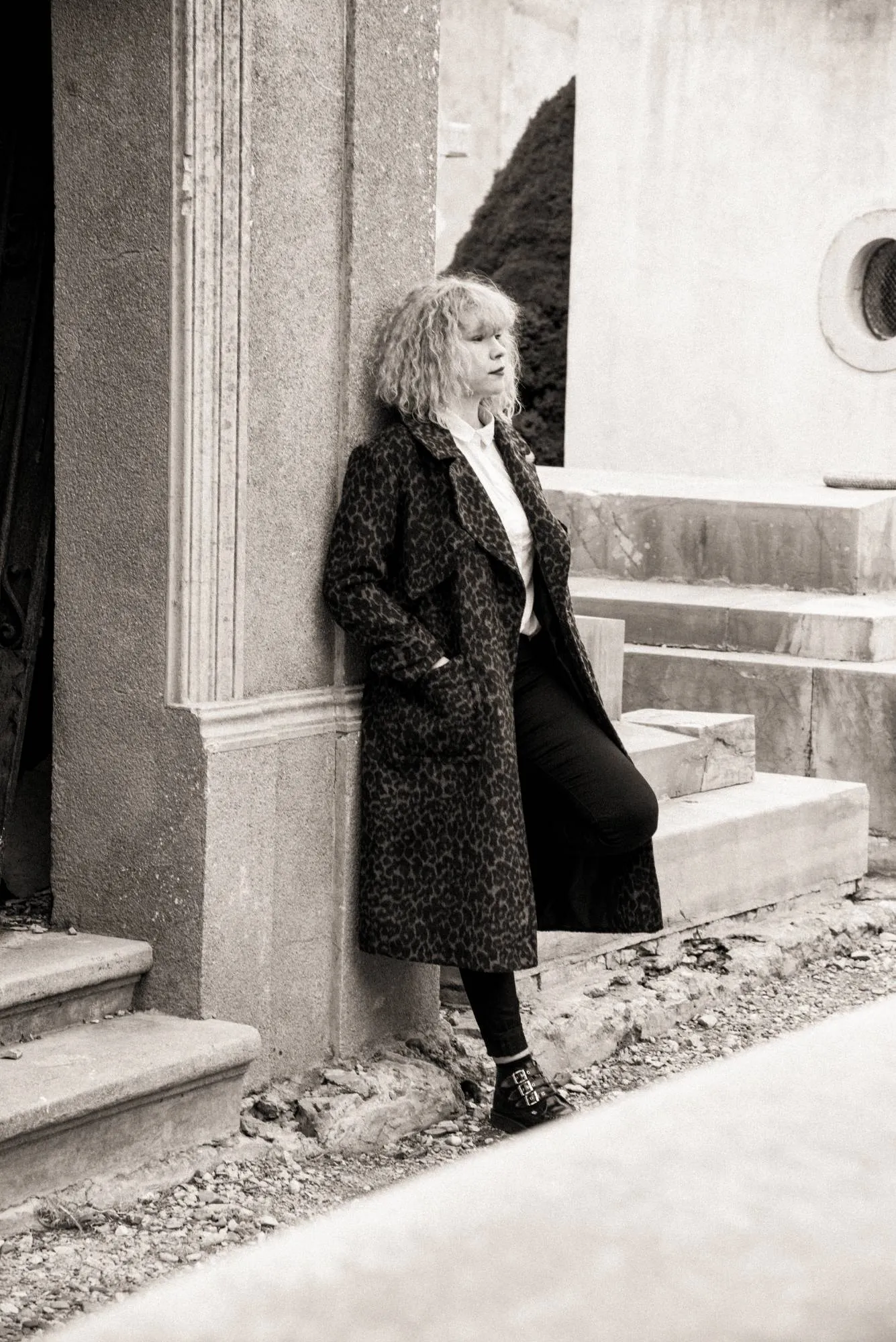 Emeraldia leaning against a stone wall, leopard coat, eyes lowered