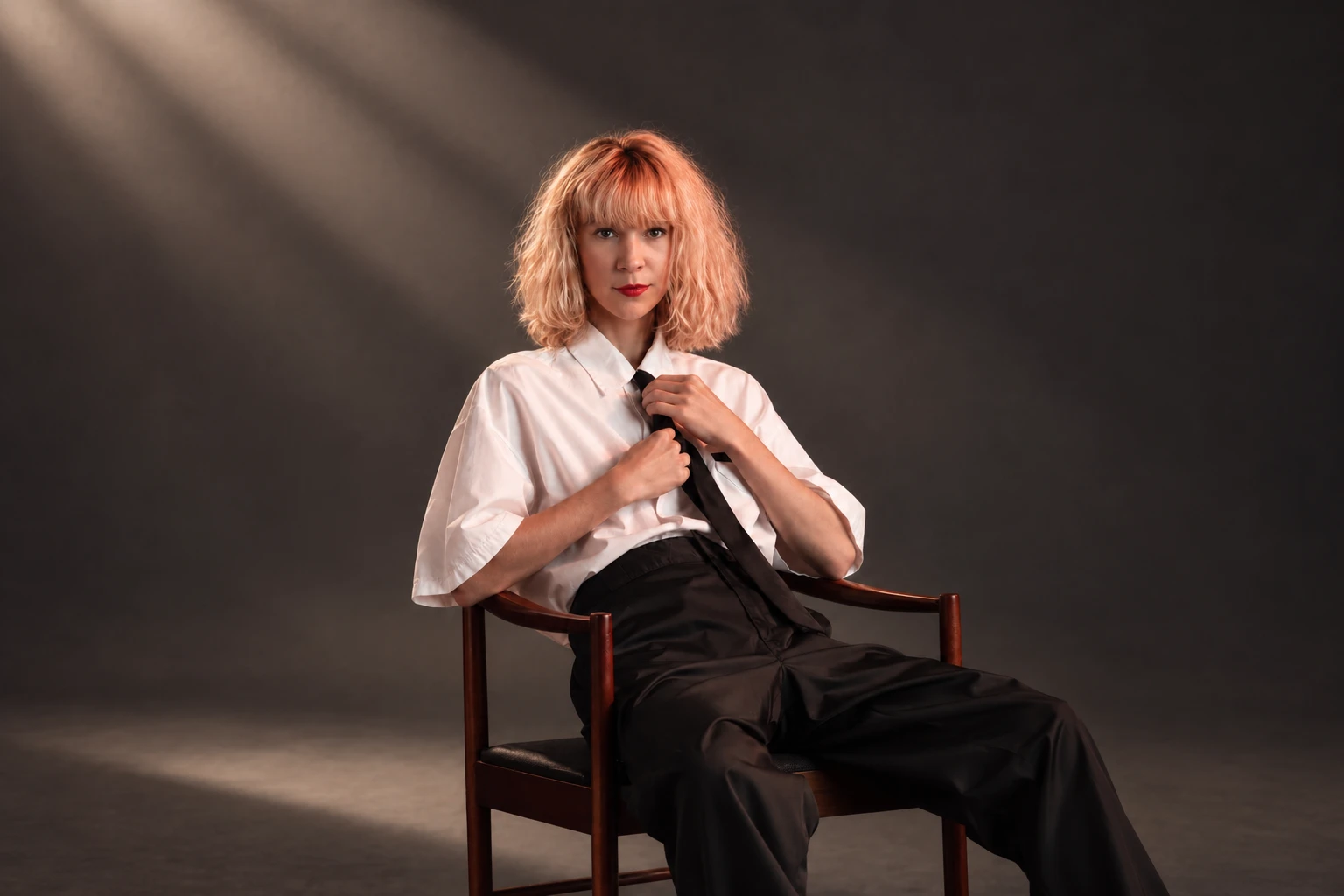 Emeraldia sitting on a wooden chair, white shirt and black tie, in a beam of light